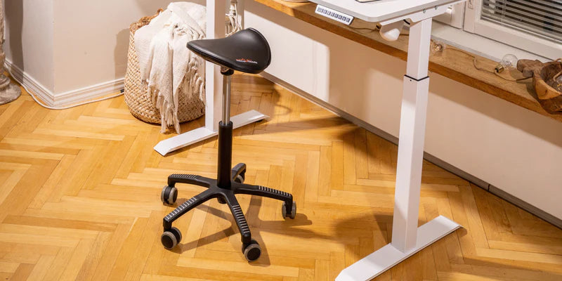 Ergonomic saddle chair with wheels next to a white adjustable desk in a home office setting.