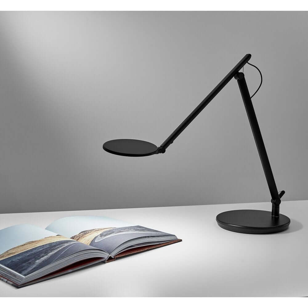 Black Nova desk lamp with sculptural design illuminates an open book on a work surface, offering glare-free, adjustable LED light.